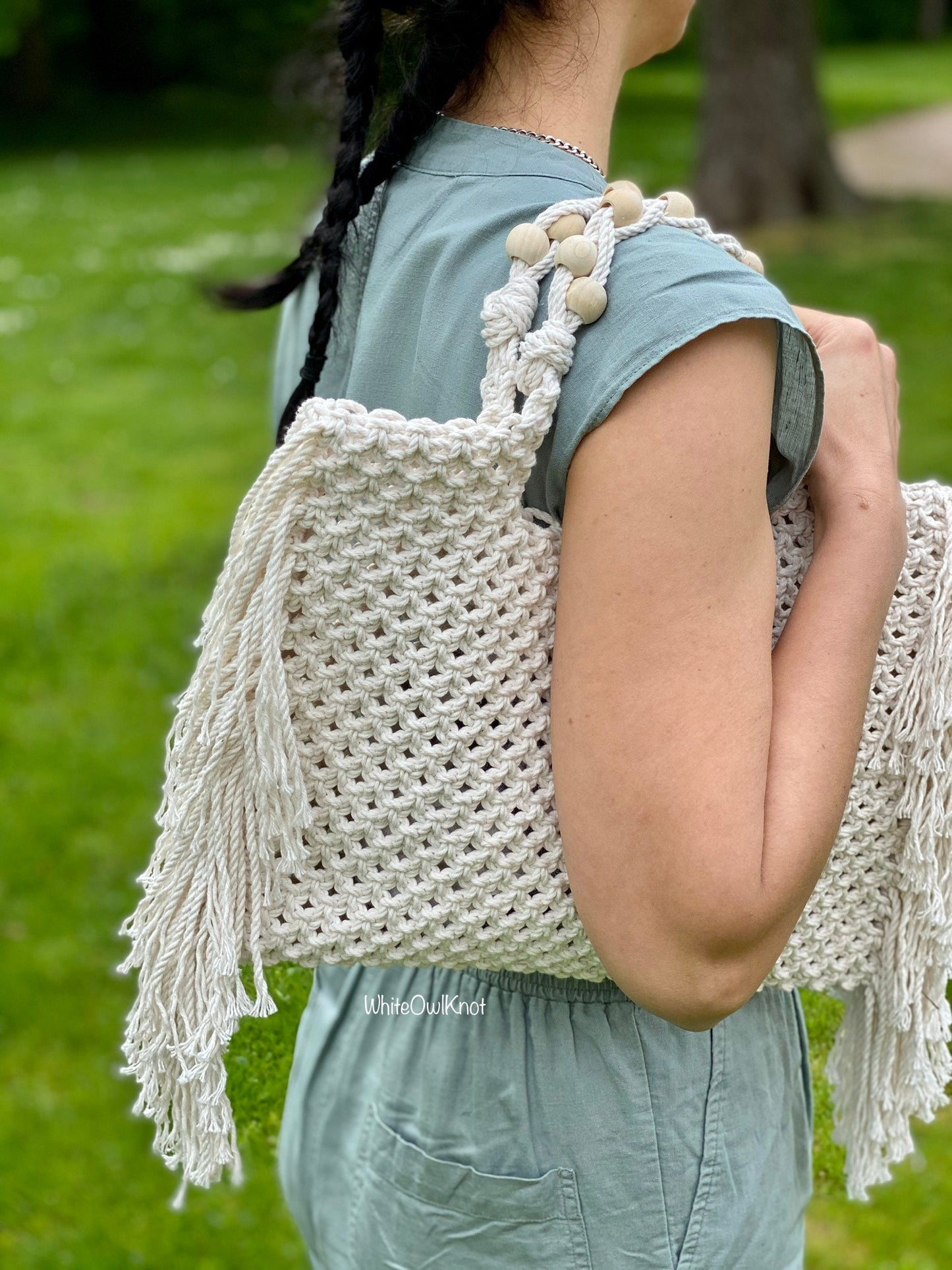Person holding a white macrame bag with tassels in a park setting