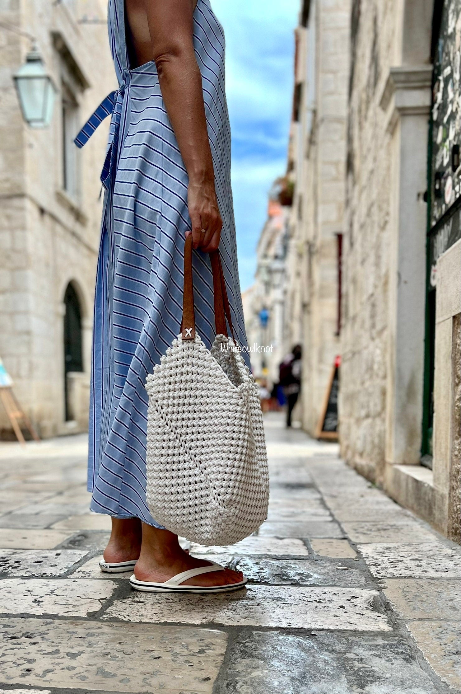 Person wearing a blue and white striped dress holding a textured handbag on a stone street.