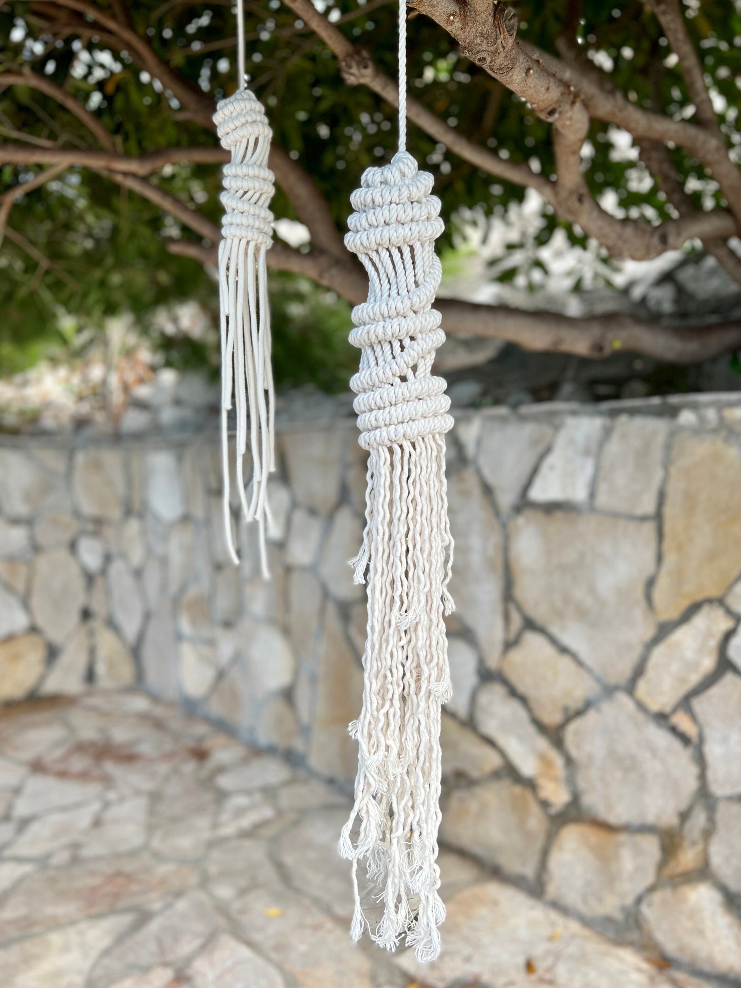 Two macrame jellyfish hangings hanging from a tree with a stone wall and greenery in the background.