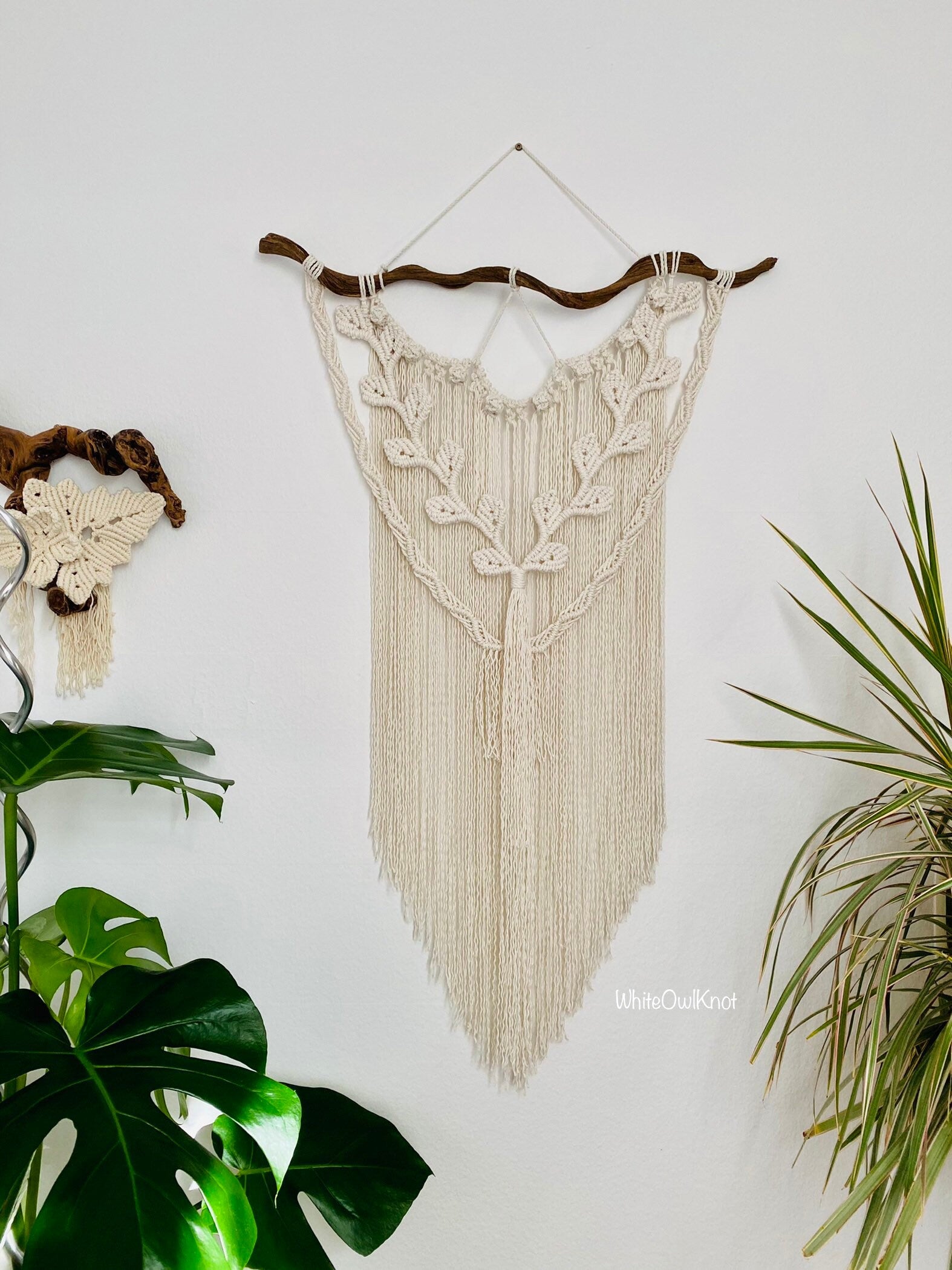 Macrame wall hanging with fringe on a white wall, surrounded by plants.