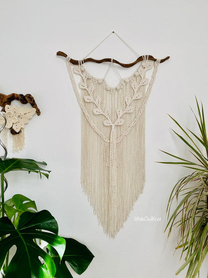 Macrame wall hanging with fringe on a white wall, surrounded by plants.