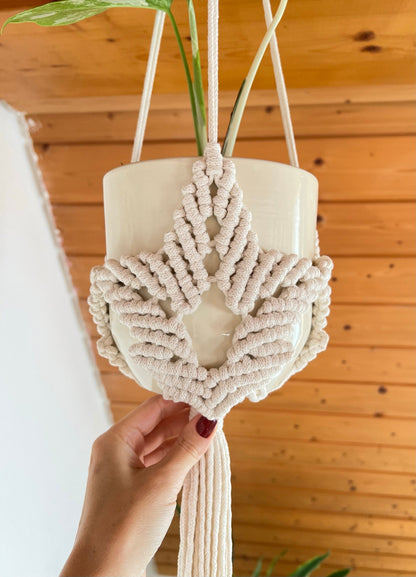 Macrame plant hanger with a heart design held by a hand against a wooden wall.