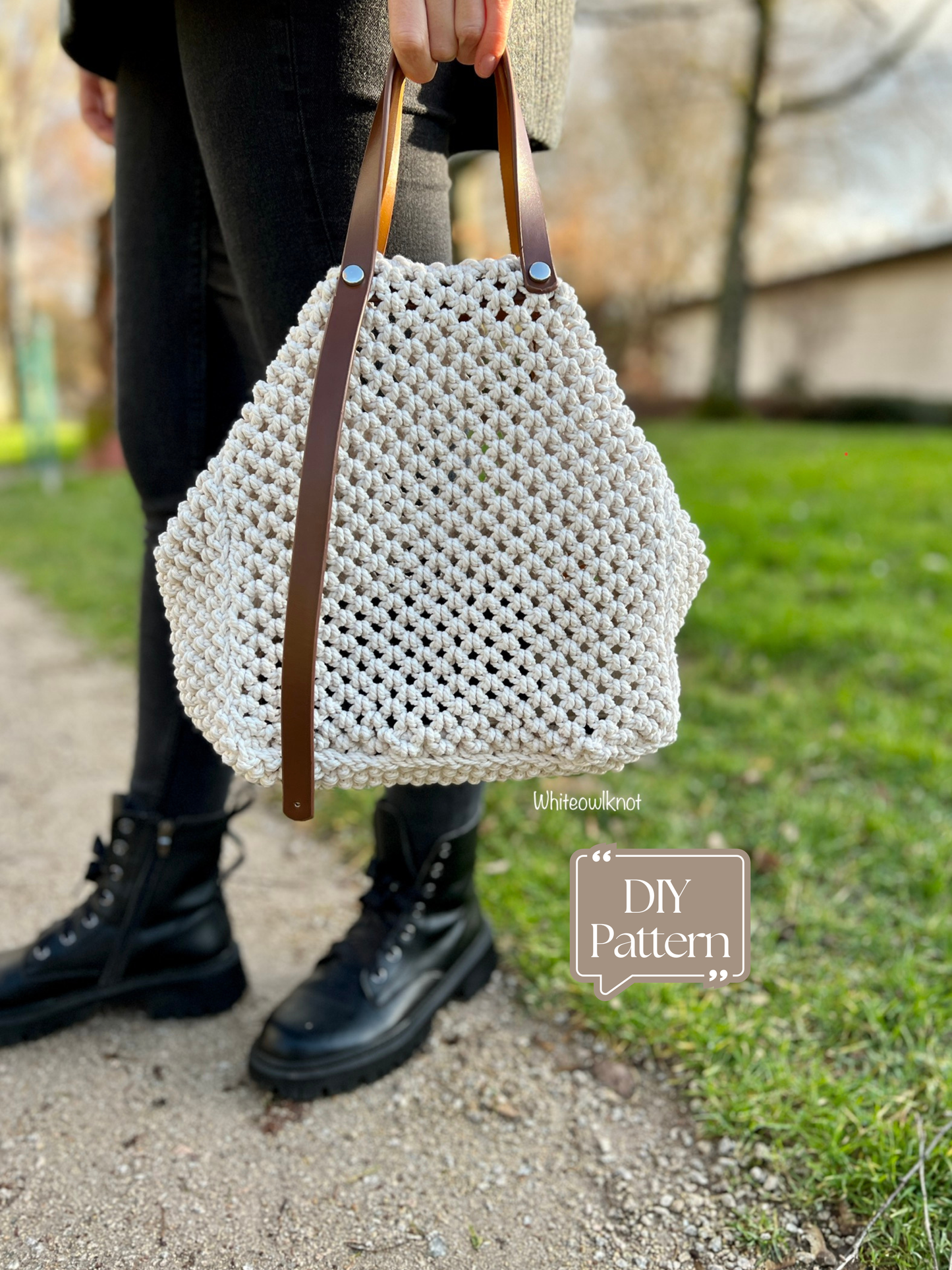 Person holding a white woven bag with brown handles on a path outdoors.
