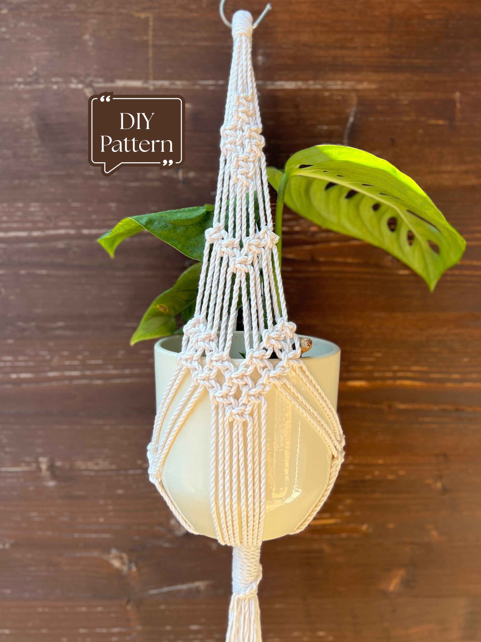 Short white macramé plant hanger holding a potted trailing plant indoors against a wooden wall; shown as a DIY macramé PDF pattern.