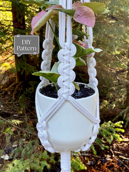 White macramé plant hanger with a braided knot detail holding a potted plant outdoors in a forest setting; shown as a DIY macramé PDF pattern.