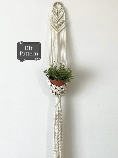 Macramé plant hanger with a long, vertical design, featuring geometric knot details at the top and a trailing fringe, holding a small terracotta pot against a white wall.