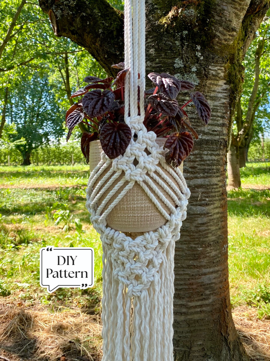 Handmade macramé plant hanger with textured geometric knot pattern, holding a fabric planter with a leafy houseplant, photographed outdoors on a tree, DIY macramé pattern example.