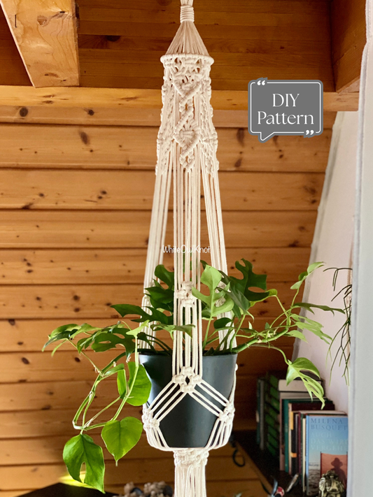White macramé plant hanger with a vintage-inspired knotted design, holding a potted houseplant indoors against a wooden wall; shown as a modern DIY macramé PDF pattern.