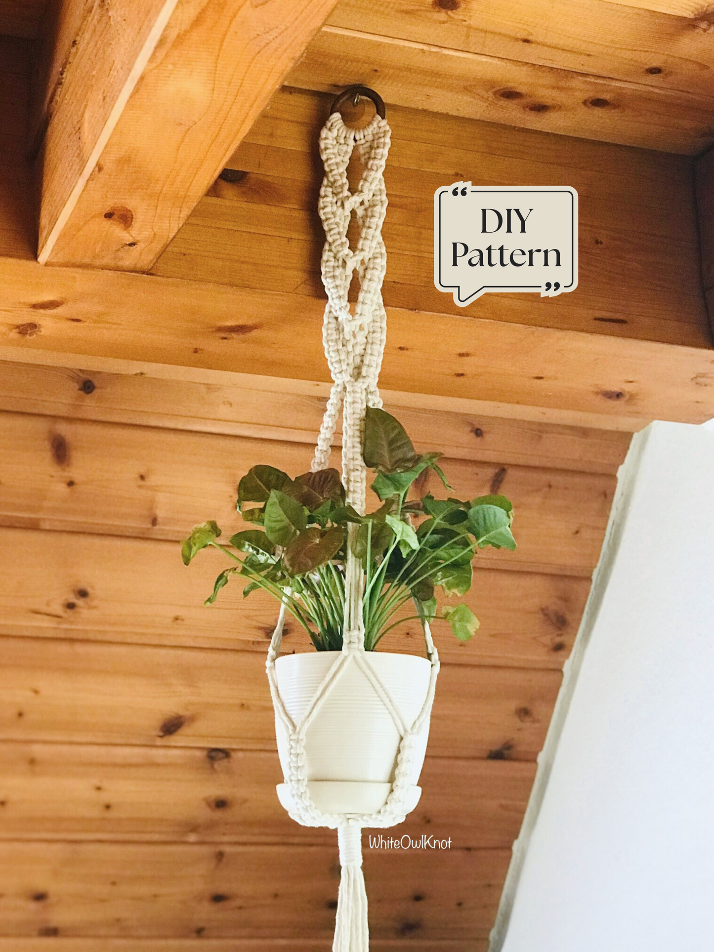Macramé plant hanger featuring an intricate decorative top knot design, supporting a white plant pot and hanging from a wooden ceiling in a warm interior setting.