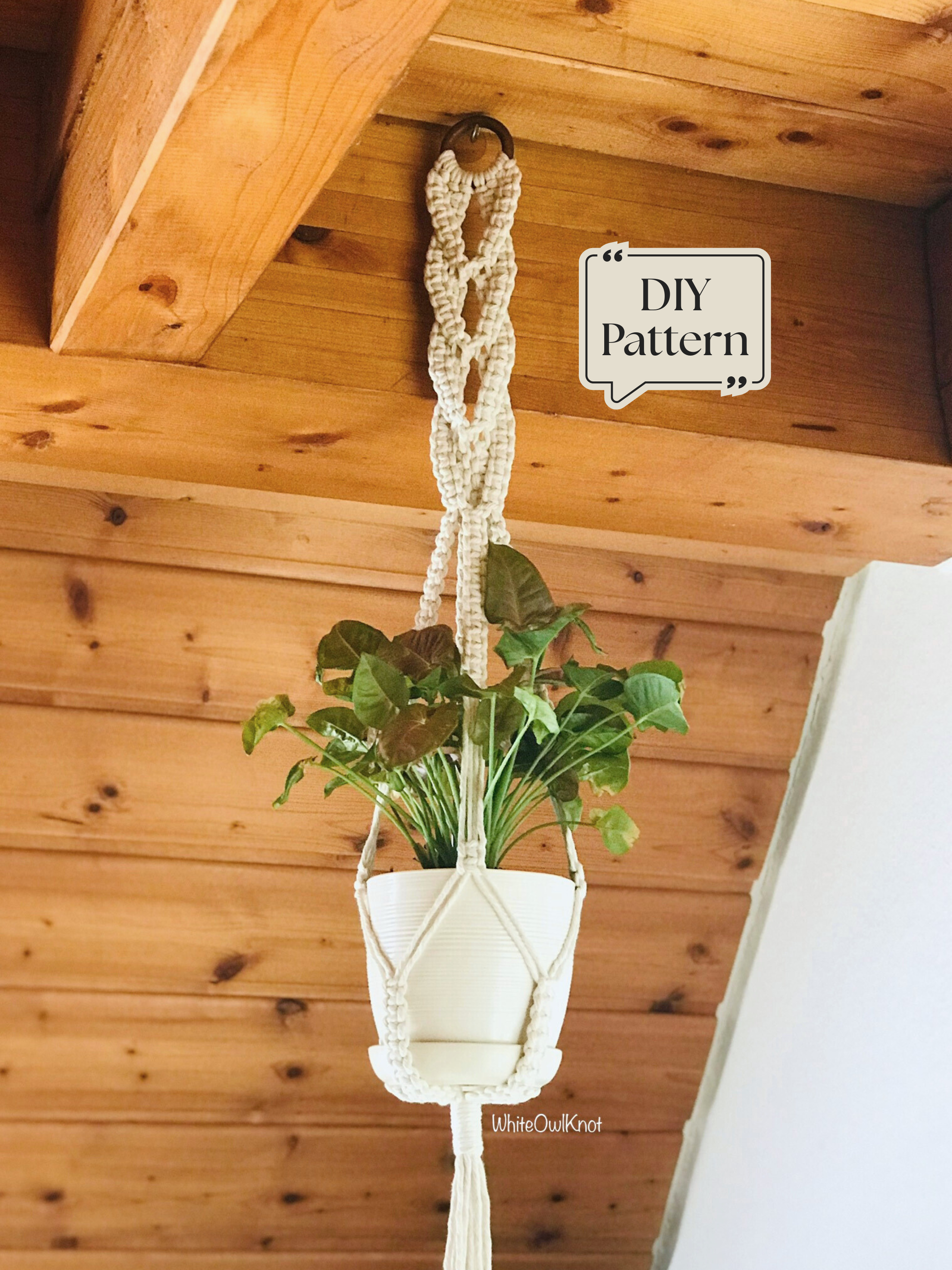 Macramé plant hanger featuring an intricate decorative top knot design, supporting a white plant pot and hanging from a wooden ceiling in a warm interior setting.