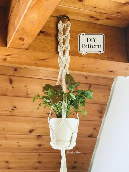 Macramé plant hanger featuring an intricate decorative top knot design, supporting a white plant pot and hanging from a wooden ceiling in a warm interior setting.