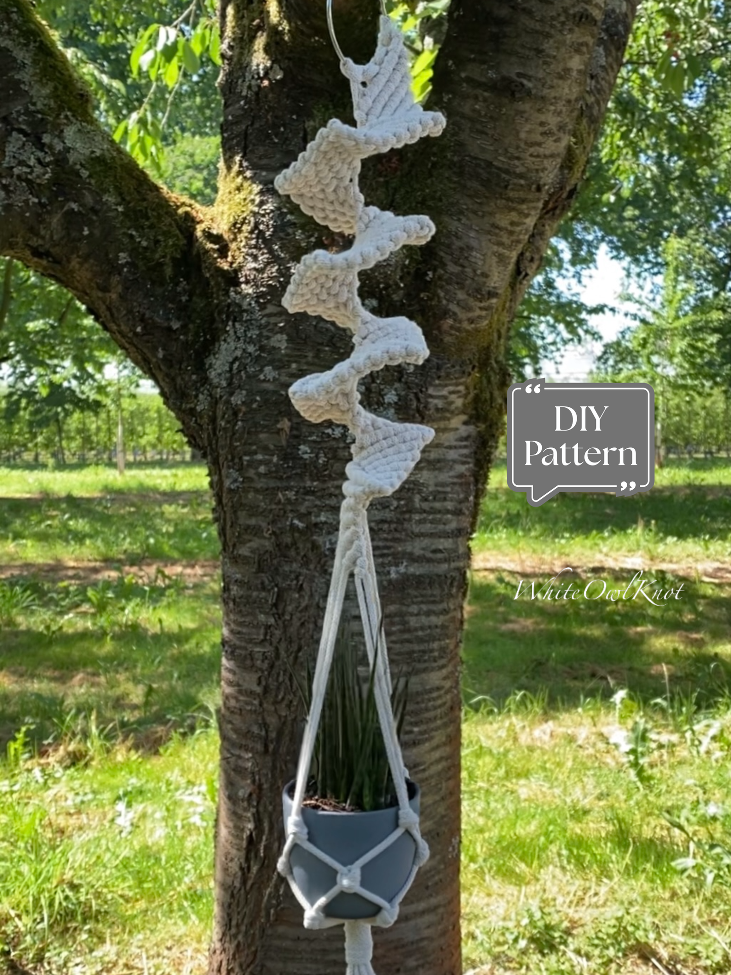 White macramé plant hanger with a sculptural spiral knot design, hanging outdoors on a tree with a potted plant; DIY macramé PDF pattern.