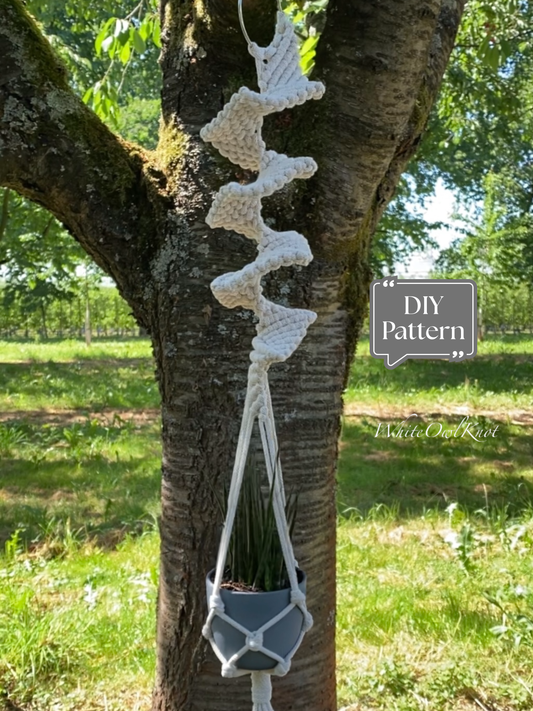 White macramé plant hanger with a sculptural spiral knot design, hanging outdoors on a tree with a potted plant; DIY macramé PDF pattern.
